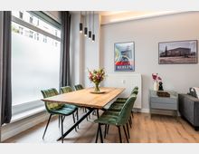 Heller, modern eingerichteter Essbereich mit großem Holztisch und mehreren grünen Polsterstühlen auf hellem Holzboden. Links ein bodentiefes Fenster mit Vorhängen, darüber hängende Pendelleuchten; an der Wand Heizkörper und gerahmte Bilder sowie ein kleines Sideboard.