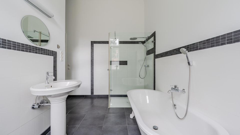 Modernes, helles Badezimmer mit freistehender Badewanne, Waschbecken mit rundem Spiegel und begehbarer Dusche mit Glasabtrennung. Weiße Wände mit dunklem Fliesenband und graue Bodenfliesen prägen den Raum.