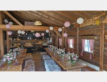 Rustikaler Innenraum einer Holzhütte mit sichtbaren Balken, langen gedeckten Holztischen und Bänken mit Fellauflagen sowie Blumenarrangements in Glasvasen. Papierlaternen hängen von der Decke, und kleine Fenster mit rot karierten Vorhängen säumen die Holzbohlenwände; im Hintergrund ist ein Barbereich zu sehen.