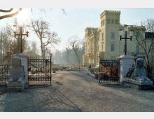 Schloss Babelsberg mit gelber Fassade und zinnenartigen Türmen, umgeben von einer Parklandschaft. Im Vordergrund eine Zufahrt mit offenem schmiedeeisernem Tor, Flammenleuchtern und steinernen Sphingen.