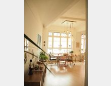 Helles Wohnzimmer mit großen, rechteckigen Fenstern, Holzboden und einem Esstisch mit Stühlen. Dekorative Elemente, Zimmerpflanzen und eine Hängelampe verleihen dem Raum eine wohnliche Atmosphäre.