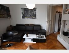 Wohnzimmer mit Parkettboden, einer schwarzen Ledercouch und einem schwarzen Sessel. Ein weißer Couchtisch steht vor der Couch, darüber hängt ein gerahmtes New-York-Bild. Rechts eine geöffnete Doppeltür, die zu einem Flur mit Schuhregal führt.