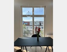 Ein heller Raum mit einem großen Fenster, das auf eine Hausfassade mit roten Dächern und Gauben blickt. Im Vordergrund steht ein schwarzer Tisch mit einer Vase roter Blumen, darüber hängen drei runde, gläserne Lampen.