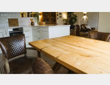 Essbereich und Küche in einem offenen Wohnraum mit einem Holztisch, gepolsterten braunen Stühlen, einer hellen weißen Küchenzeile und Dekorationselementen wie Pflanzen und Wandbildern. Im Hintergrund sind Ledersessel und warme Beleuchtung zu erkennen.