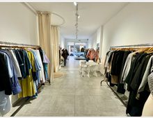 Ein minimalistisches Bekleidungsgeschäft mit Kleiderstangen auf beiden Seiten, die verschiedene Kleidungsstücke präsentieren. Im hinteren Bereich befindet sich ein kleiner Tisch mit weißen Stühlen und eine große Fensterfront, die den Blick auf die Straße freigibt.