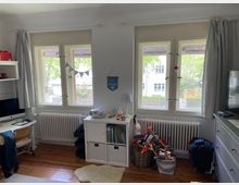 Ein Kinderzimmer mit drei Fenstern, die mit weißen Vorhängen und Rollos versehen sind. Vor den Fenstern stehen zwei Heizkörper, ein Regal mit Büchern und Spielzeug sowie Körbe mit weiteren Spielsachen. Links befindet sich ein Schreibtisch mit Computer, rechts eine Kommode und ein Fernseher. Der Boden ist aus Holz, teilweise mit einem Teppich bedeckt.