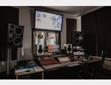 Tonstudio mit einem großen Mischpult voller Regler und beleuchteter Anzeigen in der Mitte, umgeben von mehreren Lautsprechern. Im Hintergrund ein Fenster mit Blick auf einen Aufnahmeraum und darüber ein Monitor, der das Bild des Studios zeigt.