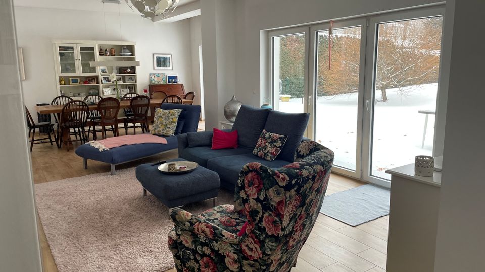 Offener Wohn- und Essbereich mit blauem Ecksofa, Hocker und gemustertem Sessel auf Teppich sowie einem langen Esstisch mit Stühlen und Vitrinenschrank im Hintergrund. Große bodentiefe Glas-Schiebetüren führen in einen verschneiten Garten mit Bäumen.