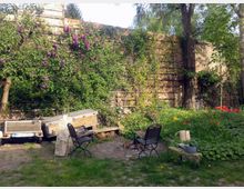 Ein kleiner, grüner Garten mit einer alten Backsteinmauer, die von Kletterpflanzen überwuchert ist. Es gibt blühende Blumen, zwei Metallstühle, eine Gießkanne, einen Baum sowie einen abgestellten Anhänger im Hintergrund.