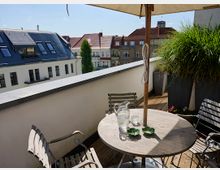 Dachterrasse mit Holzboden, einem runden Holztisch und Metallstühlen. Auf dem Tisch stehen eine Glaskaraffe, Gläser und Schalen; ein Sonnenschirm spendet Schatten. Im Hintergrund sind Wohnhäuser mit roten Dächern und Fensterfronten zu sehen.