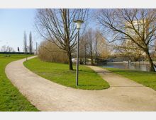 Ein Park mit grünen Rasenflächen, geschwungenen Kieswegen und mehreren laubfreien Bäumen. Im Hintergrund ist ein Fluss mit einer Brücke und einem großen Gebäude sichtbar, außerdem eine Laterne und einige Bänke entlang der Wege.