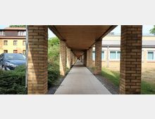 Langer überdachter Gehweg mit hellen Backsteinpfeilern und Holzdecke, flankiert von Grünflächen und Gebäuden mit gelben Fassaden und Fenstern. Im Hintergrund ein geparktes Auto und weitere Gebäude.