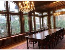 Historischer Speisesaal mit großen Fenstern aus verziertem Buntglas, einem langen Holzesstisch und passenden Stühlen. Zwei Kronleuchter hängen von einer Holzdecke, und draußen sind Bäume sichtbar.