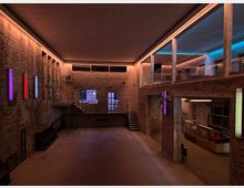 Industriehalle mit modernem Innenausbau: Der Raum hat unverputzte Backsteinwände, hohe Decken und eine Kombination aus farbiger LED-Beleuchtung sowie Oberlichtern. Eine schmale Treppe führt zu einer verglasten Galerie im oberen Bereich, und auf der unteren Ebene sind Sitzgelegenheiten sowie Vitrinen sichtbar.