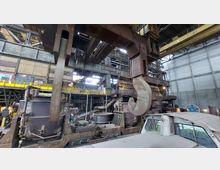 Innenraum einer großen Industriehalle mit metallischen Strukturen, einem massiven Kranhaken im Vordergrund und schweren Maschinen wie Schmelzöfen und Förderanlagen im Hintergrund. Die Umgebung wird durch Stahlträger, Ziegelwände und Geländer aus gelbem Metall definiert.