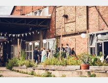 Backsteinfassade eines Gebäudes mit großen Fenstern, davor eine Gruppe elegant gekleideter Menschen im Gespräch. Der Bereich ist mit Hochbeeten, Pflanzen und hängenden Lichterketten sowie Wimpelgirlanden dekoriert.
