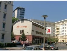 Straßenszene mit einem Gebäude im Vordergrund, das den Schriftzug „feuersozietät“ trägt. Das Gebäude hat eine gewölbte Dachform und ist von Bäumen, geparkten Autos sowie weiteren mehrstöckigen Häusern im Hintergrund umgeben.