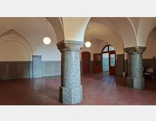Gewölbte Eingangshalle mit großen Granitsäulen und Rundbogen-Decke, hängenden Kugelleuchten sowie rotbraunem Boden und grauer Wandverkleidung. Im Hintergrund befinden sich Holztüren mit Glaseinsätzen und ein hoher Rundbogen-Durchgang.