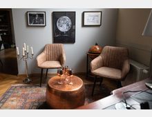 Ein stilvoll eingerichteter Raum mit zwei braunen Polstersesseln, einem runden kupferfarbenen Tisch mit drei metallischen Weingläsern und einem Teppich mit orientalischem Muster. An der Wand hängen Bilder, darunter eine Darstellung des Mondes, und im Hintergrund steht ein Globus auf einem kleinen Holzständer.