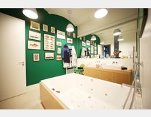 Modernes Badezimmer mit grüner Akzentwand, dekoriert mit gerahmten Illustrationen von Fischen und Schiffen. Im Raum befinden sich eine große Badewanne, ein Doppelwaschbecken mit Holz-Unterschrank und ein großer Spiegel, beleuchtet durch mehrere Hängelampen.