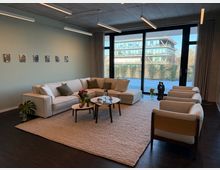 Ein modern eingerichtetes Wohnzimmer mit einer hellen Eckcouch, drei hellen Sesseln und einem runden Couchtisch auf einem beigefarbenen Teppich. Im Hintergrund ist eine große Fensterfront mit Blick auf einen begrünten Außenbereich und ein modernes Bürogebäude.