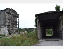 Verlassene Industrieanlage mit einem zerfallenen Betonsilo links und einer offenen, tunnelartigen Betonstruktur rechts. Im Hintergrund sind überwucherte Vegetation und Baumlandschaft zu sehen.