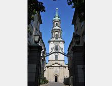 Barocke Kirchenfassade mit einem hohen, kunstvoll verzierten Turm und einer goldenen Spitze, flankiert von symmetrischen Gebäuden. Ein geschwungenes schmiedeeisernes Tor mit Ornamenten überspannt den Eingangsbereich.