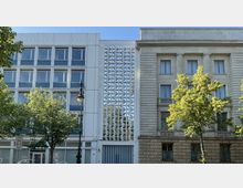 Moderne Gebäudeansicht mit drei Fassadenteilen: links ein weißes Gebäude mit quadratischen Fenstern, in der Mitte ein Element mit durchbrochenem Betonmuster, und rechts ein historisch anmutendes Gebäude mit Säulen und Steinverkleidung. Im Vordergrund stehen Bäume und eine klassische Straßenlaterne.