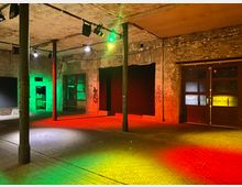 Industrieähnlicher Innenraum mit unverputzten Backsteinwänden, sichtbaren Metallstützen und teils vergitterten Fenstern. Der Raum ist in roten, gelben und grünen Lichtern beleuchtet, mit Graffiti an einigen Wänden und einer großen Doppeltür aus Holz und Glas rechts im Bild.