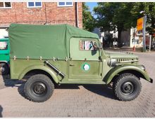 Ein olivgrüner Geländewagen mit Planenabdeckung steht auf einem gepflasterten Platz, umgeben von einem roten Backsteinbau im Hintergrund und Bäumen. Rechts im Bild sind ein Netto-Supermarkt-Schild und weitere Gebäude sichtbar.