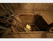 Blick in einen alten Kellerraum mit grob gemauerten Steinwänden. Eine Holzleiter führt hinunter, Rohre und eine Seilwinde sind sichtbar, die Umgebung wirkt rustikal und unverputzt.