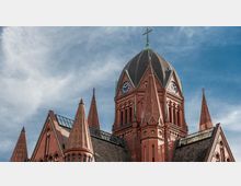 Außenansicht der Turmspitze einer neugotischen Backsteinkirche mit mehreren spitzen Türmen, runden Fenstern und einer großen Kuppel mit Kreuz an der Spitze; zwei Uhren sind an der Kuppel angebracht, der Himmel ist klar und leicht bewölkt.