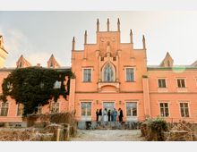 Rosafarbenes, schlossähnliches Gebäude im neugotischen Stil mit zentralem Giebel, schlanken Ziertürmchen und Spitzbogenfenster über dem Eingang. Links bedeckt dichter Efeu einen großen Teil der Fassade; davor liegt ein Gartenbereich mit Beeten und Wegen.
