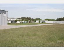 Flugplatzgelände mit einer großen Grünfläche im Vordergrund, einem asphaltierten Weg und mehreren flachen, weißen Industriegebäuden im Hintergrund. Im hinteren Bereich sind vereinzelt Bäume und Sträucher zu sehen.