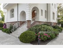 Eingangsbereich eines weißen Wohngebäudes mit Ziegelbasis, geschwungenen Treppengeländern aus Metall und einem hohen, bogenförmigen Türrahmen. Umgeben von gepflegten Sträuchern, blühenden Hortensien und einer gepflasterten Terrasse.