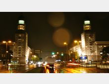 Nachtaufnahme einer breiten Straße in einer Stadt, flankiert von zwei symmetrischen Gebäuden mit beleuchteten Türmen. Die Straße ist nass von Regen, und Lichter von Straßenlaternen, Verkehrsampeln und vorbeifahrenden Autos spiegeln sich auf dem Asphalt.