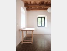 Ein heller Innenraum mit glatten, beigen Wänden, einem Boden aus poliertem Stein und einer Holzdecke mit sichtbaren Balken. Im Raum stehen ein einfacher Holzwerktisch und zwei Fenster mit schwarzen Rahmen, die natürliches Licht hereinlassen.