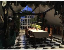 Ein rustikaler Innenraum mit einem langen Esstisch, bedeckt von einer Tischdecke und dekoriert mit Pflanzen in Töpfen, umgeben von Holzstühlen. Der Boden ist schwarz-weiß kariert, und im Hintergrund befindet sich ein großes Fenster mit Lichterketten und Blick auf dunkle, blaue Abendstimmung; der Raum ist mit Cowboyhüten, Sätteln und Pflanzen dekoriert.