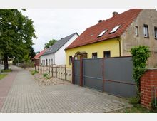 Straßenszene mit Einfamilienhäusern, darunter ein gelbes Haus mit rotem Ziegeldach und eingezäuntem Vorgarten. Ein gepflasterter Gehweg verläuft entlang der Häuser, gesäumt von Bäumen und einem gemischten Gartenbereich.