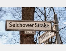 Straßenschilder mit den Aufschriften „Selchower Straße“ und „Lichtenrader Straße“, montiert an einem Metallpfosten vor einem Hintergrund mit kahlen Baumästen und blauem Himmel.