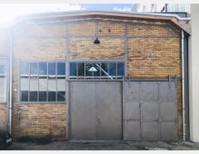 Industrielle Backsteinfassade mit großen Metalltüren und einem Dach aus Metallträgern. Oberhalb der Türen befinden sich Fenster aus Glas und Metallrahmen, die symmetrisch angeordnet sind.