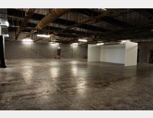 Große, leere Industriehalle mit Betonboden und grauen Steinwänden, beleuchtet von langen Deckenleuchten. An der offenen Decke sind sichtbare Rohre, Kabeltrassen und Lüftungsleitungen, rechts steht eine freistehende weiße Trennwand.