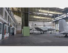Innenansicht eines historischen Flugzeughangars mit einem silbernen Propellerflugzeug, das zur Ausstellung steht. Der Hangar hat weiße Backsteinwände, große Fenster, eine Metallträgerdecke und industrielle Beleuchtung.