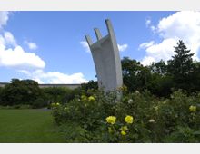 Ein modernes Denkmal aus Beton mit gebogenen Säulen ragt hoch in den Himmel. Es steht inmitten eines Parks, umgeben von gelben Rosen und dichtem grünen Laub, unter einem blauen Himmel mit weißen Wolken.