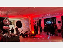 Ein moderner Innenraum mit Partyatmosphäre, beleuchtet durch rotes Licht und Disco-Kugeln, die Lichtreflexionen erzeugen. Im Vordergrund befindet sich eine Bar mit Getränken, im Hintergrund Menschen in Gesprächen sowie eine Leinwand mit einer Projektion.