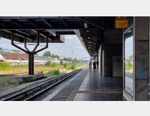 Bahnsteig eines Bahnhofs mit teils überdachtem Bereich, Gleisen auf der linken Seite und einer Fußgängergruppe rechts. Im Hintergrund sind Bahngleise, Züge, ein Gebäude und ein gestaffeltes Stadtpanorama sichtbar.