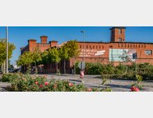 Backstein-Gebäude mit historischem Industriebau-Charakter, genutzt als Gasthaus, Hotel und Freizeitstätte, mit Werbeschildern an der Fassade. Im Vordergrund eine mit Rosen und Bäumen bepflanzte Straße unter blauem Himmel.