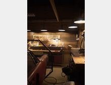 Innenansicht einer Werkstatt mit Holzvertäfelung, großer Werkbank und einer Werkzeugwand mit aufgehängten Schraubenschlüsseln und Zangen. Mehrere Pendelleuchten hängen von der dunklen Decke, daneben stehen Werkstattgeräte und links ist ein teilweise sichtbares Fahrzeug oder Maschinengehäuse.