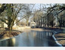 Ein zugefrorener Teich in einer Parklandschaft mit kahlen Bäumen im Winter. Im Hintergrund ist ein kleines Gebäude mit Säulen und Satteldach zu sehen, das von Bäumen umgeben ist.