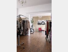 Heller, loftartiger Boutique- oder Showroomraum mit Holzboden, Kleiderständern voller glitzernder Kleidung und Taschen sowie Kronleuchtern an der Decke. Im hinteren Bereich stehen rote Sessel vor einer beige strukturierten Wand mit drei gerahmten Bildern; daneben ein Fahrrad und eine Topfpflanze.